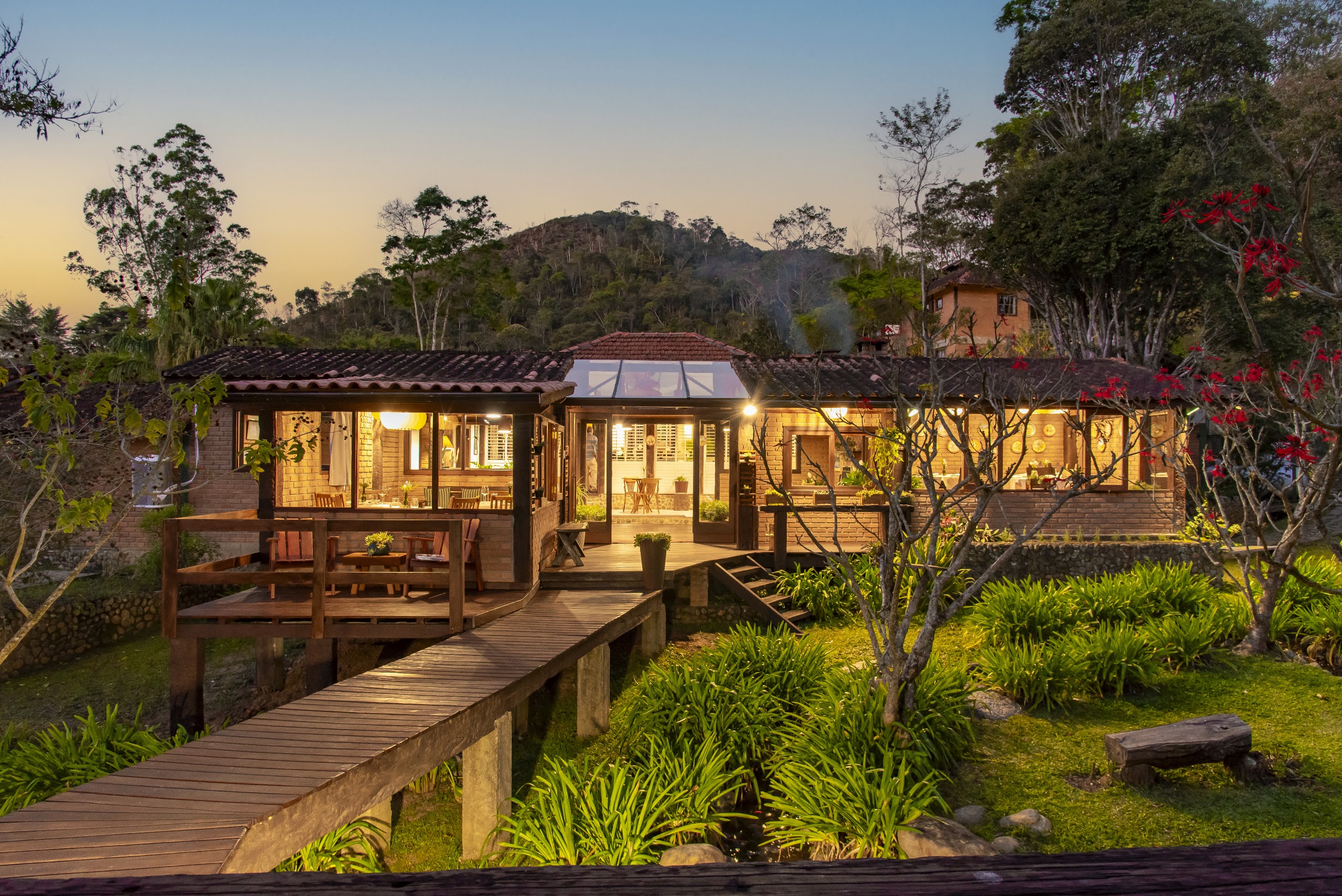 Restaurante iluminado ao anoitecer, com passarela de madeira e a serra ao fundo
